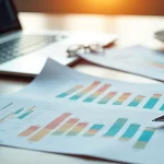 Mesa de escritório com gráficos financeiros impressos, calculadora e laptop aberta exibindo gráficos de investimentos